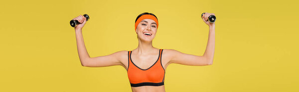 cheerful woman in orange sports bra working out with dumbbells isolated on yellow, banner