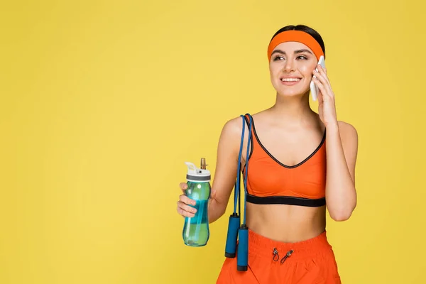 スポーツボトルとジャンプロープを持つ笑顔の女性は黄色に隔離されたスマートフォンを呼び出し — ストック写真