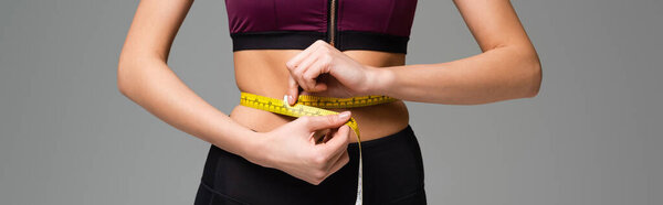 partial view of sportswoman measuring waist isolated on grey, banner
