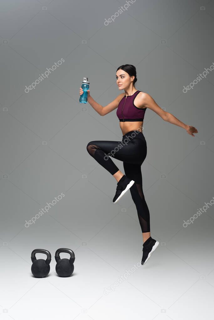 young woman with sports bottle training near kettlebells on grey background