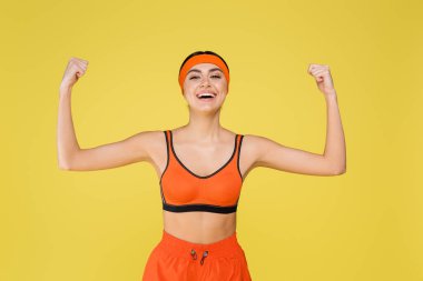 cheerful sportswoman demonstrating strength while smiling at camera isolated on yellow clipart