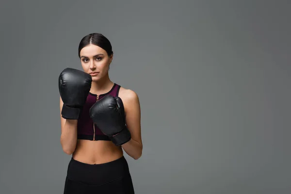slim woman in black sportswear and boxing gloves looking at camera isolated on grey