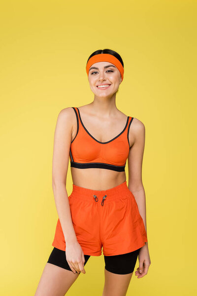 young woman in orange sportswear smiling at camera isolated on yellow