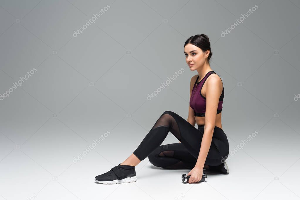 young sportswoman looking away while sitting with dumbbell on grey background