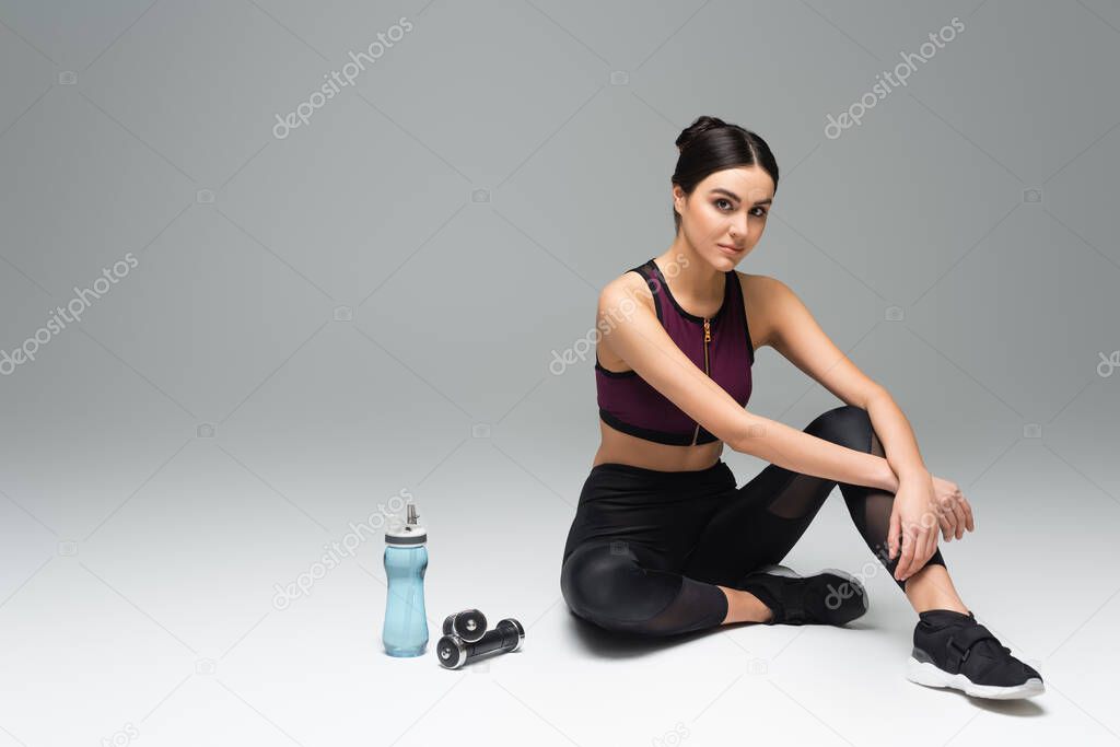 sportswoman sitting near sports bottle and dumbbells while looking at camera on grey background