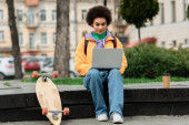 Afroamerikanische Freelancer mit Laptop in der Nähe von Coffee to go und Longboard im Freien 