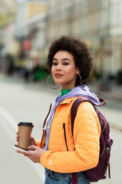 Sırt çantalı, kağıt bardaklı ve akıllı telefonlu genç Afro-Amerikan kadın dışarıda kameraya bakıyor. 