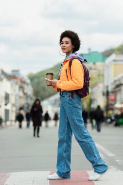 Afro-Amerikalı kadın elinde kahveyle şehir caddesinde akıllı telefonuyla dolaşıyor. 
