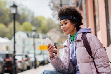 Şehir sokağındaki bulanık binanın yakınında akıllı telefonu olan Afrikalı Amerikalı kadın. 
