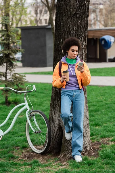 Mujer Afroamericana Con Café Para Llevar Smartphone Pie Cerca Árbol — Foto de Stock