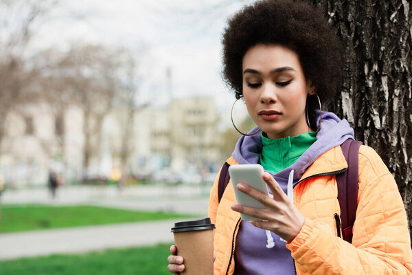 Pretty african american woman in jacket using cellphone and holding coffee to go near tree 