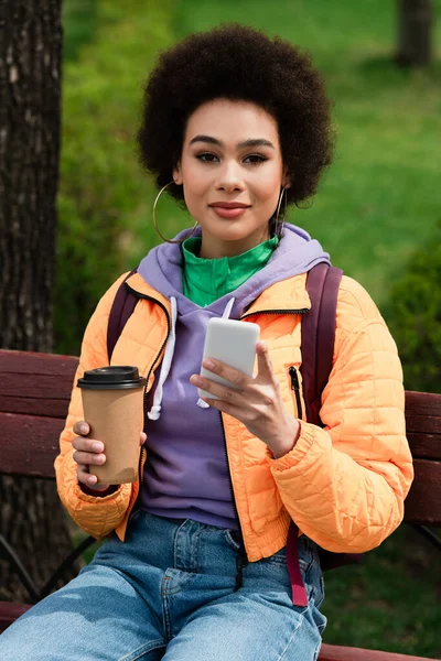 Mujer Afroamericana Con Teléfono Celular Taza Papel Mirando Cámara Banco —  Fotos de Stock