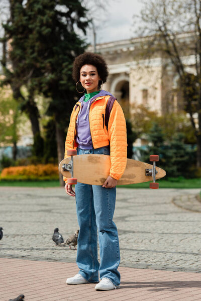 Африканская американка с длинной доской смотрит в камеру рядом с размытыми голубями на улице 