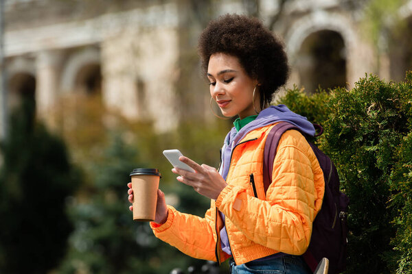 Pretty african american woman holding coffee cup and using cellphone near bush outdoors 