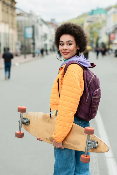 Uśmiechnięta Afroamerykanka Plecakiem Longboardem Stojąca Miejskiej Ulicy — Zdjęcie stockowe