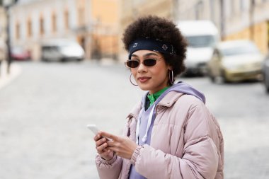 Güneş gözlüklü ve bandanalı Afro-Amerikalı kadın şehir caddesinde cep telefonu tutuyor. 