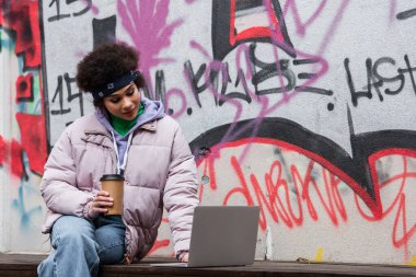 Afro-Amerikalı bir kadın elinde kahve tutup dışarıda grafiti yapmak için dizüstü bilgisayar kullanıyor. 