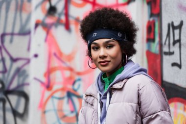 Afro-Amerikalı bir kadın dışarıda bulanık grafitilerin yakınındaki kameraya bakıyor. 