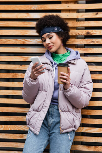 African american woman with coffee to go using cellphone near wooden fence 