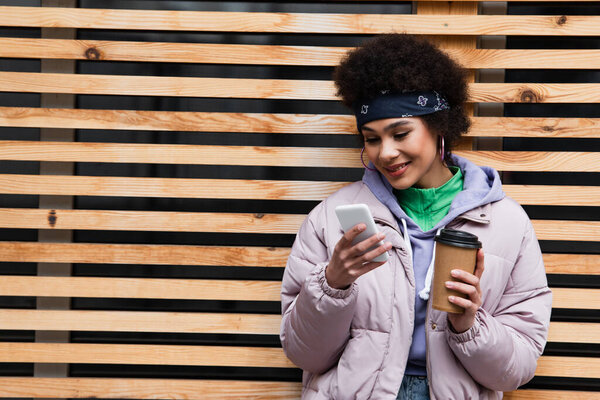 Positive african american woman using mobile phone and holding paper cup near fence 