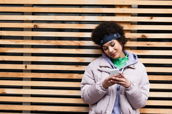 木製のフェンスの近くに立っている間 スマートフォンを使用してスタイリッシュなアフリカ系アメリカ人女性 — ストック写真