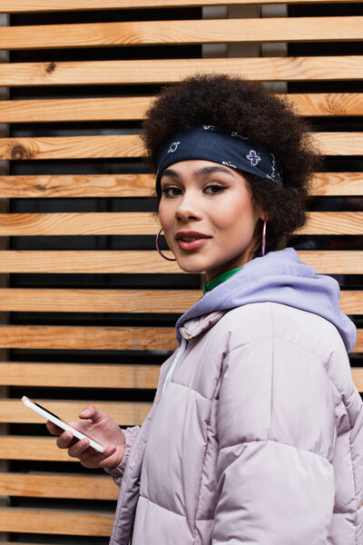 African american woman in jacket holding mobile phone and looking at camera outdoors 
