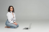 Svůdná žena v bílé košili při pohledu na kameru v blízkosti notebooku na šedém pozadí 