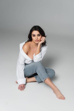 Çıplak ayaklı kadın gri arka planda otururken kameraya bakıyor.