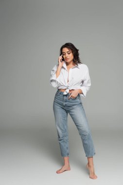 Çıplak ayaklı kadın gri arka planda akıllı telefondan konuşuyor.