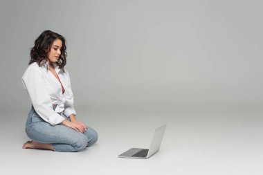 Gri arka planda otururken dizüstü bilgisayara bakan seksi esmer kadın. 