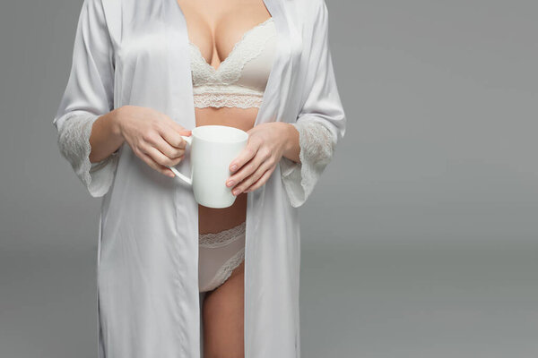 Cropped view of woman in underwear and satin robe holding white cup isolated on grey