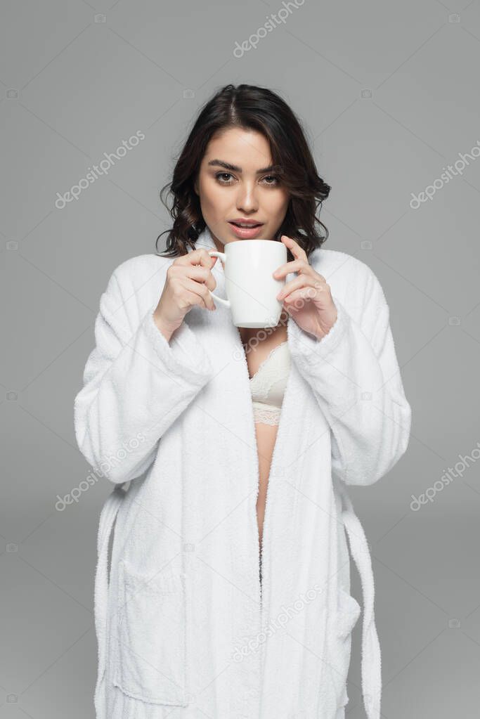 Pretty woman in bra and bathrobe holding cup isolated on grey