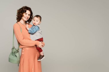 Saten etekli mutlu kadın kollarında yürümeye yeni başlamış oğlu gri üzerinde izole edilmiş.