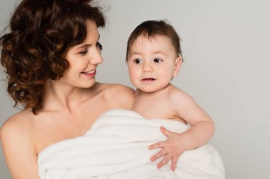 Gülümseyen bir anne, omuzlarında küçük bir çocukla gri bir havluya sarılmış bir şekilde.