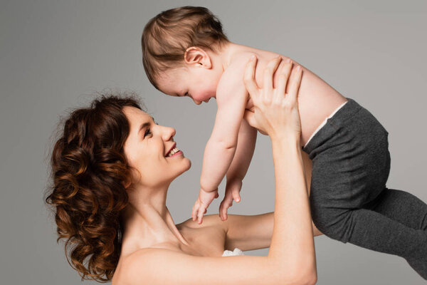 happy mother with bare shoulders holding in arms son in baby tights isolated on grey