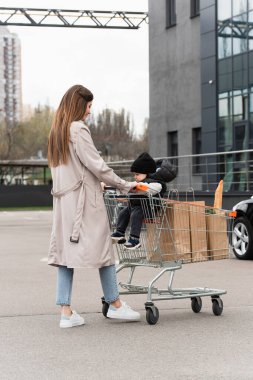 Trençkotlu genç bir kadın oğluyla yürüyor ve alışveriş arabasıyla araba parkında alışveriş yapıyor.