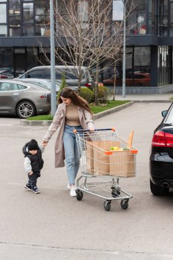 Araba parkında alışveriş arabasıyla yürürken çocuğun elini tutan genç bir kadın.