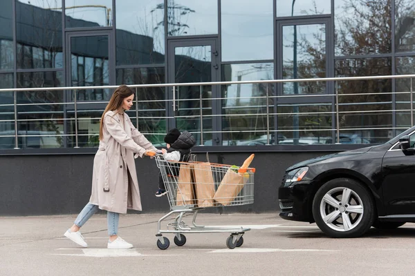 息子を持つ若い女性と車に向かって歩くショッピングカートで購入 — ストック写真