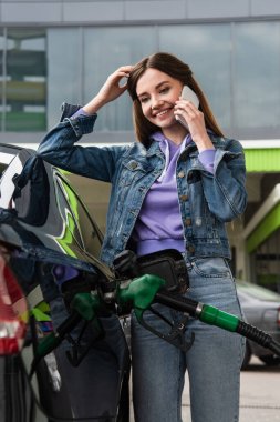 cheerful woman talking on mobile phone while refilling car on gas station clipart