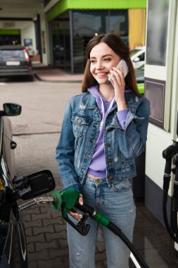 Kot elbiseli neşeli kadın benzin istasyonunda cep telefonuyla konuşuyor.