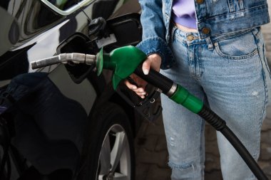 Benzin istasyonunda, arabanın yanında elinde yakıt tabancası olan kot pantolonlu bir kadın görülüyor.