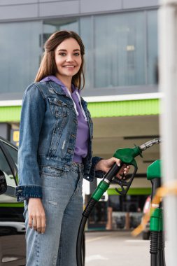 happy woman in denim jacket holding fuel pistol on gas station clipart