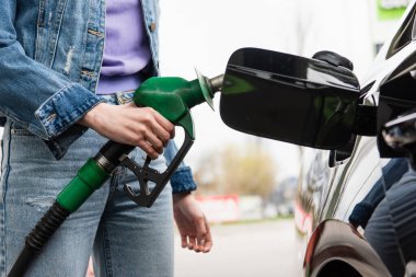 partial view of young woman in jeans holding gasoline pistol near automobile clipart
