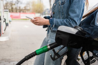 partial view of young woman messaging on cellphone near car on gas station clipart