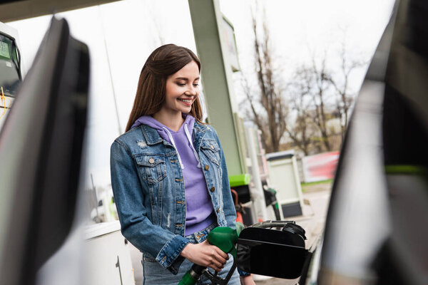 happy woman in denim jacket refueling blurred car on gas station