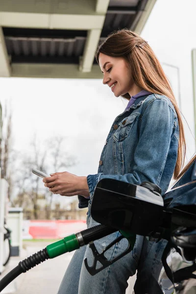 ガソリンスタンドで自動車を給油しながら携帯電話でメッセージを送るかなりの女性は — ストック写真