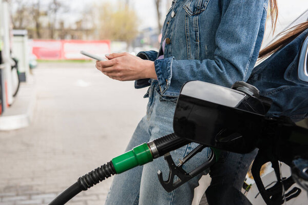 partial view of young woman messaging on cellphone near car on gas station