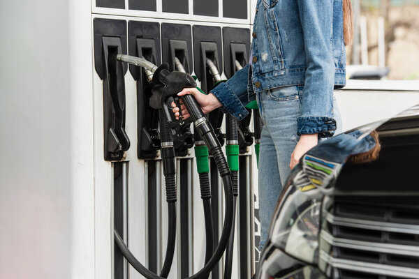 partial view of woman in denim clothes near fuel pistols and blurred car on gas station