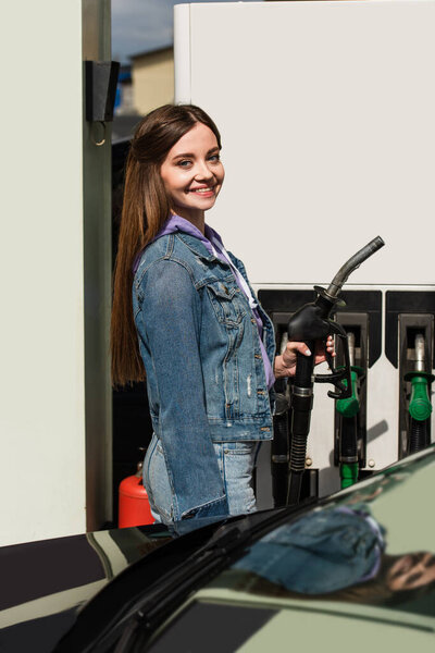 happy young woman smiling at camera while holding fuel pistol on gasoline station