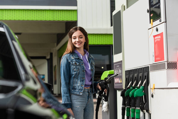 smiling woman in denim clothes holding gasoline pistol near blurred car